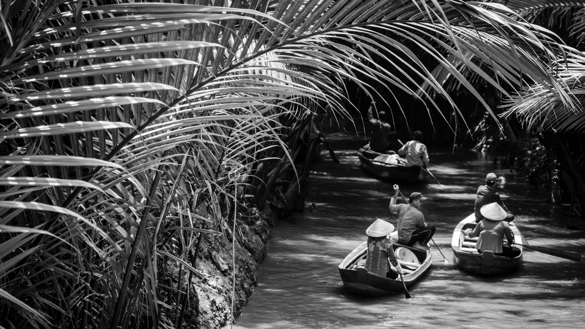 Black and white photo of small traditional boats navigating a jungle river with tropical foliage.