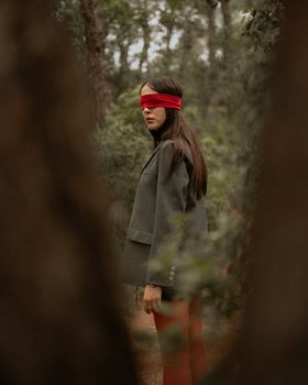 Artistic photo of a woman in a gray suit and red blindfold standing in a forest.