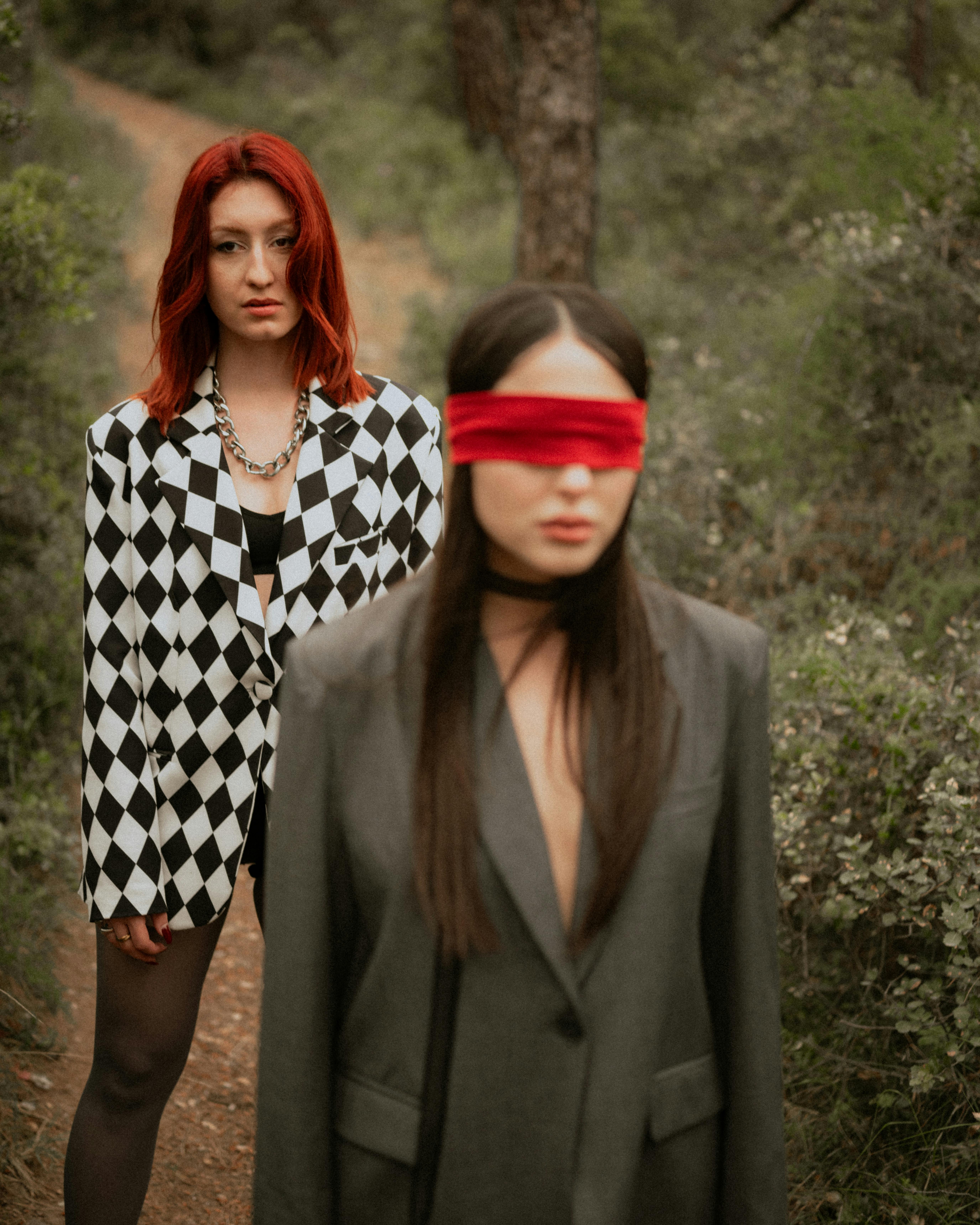 Two women in stylish outfits, one blindfolded, posing mysteriously in an outdoor forest.