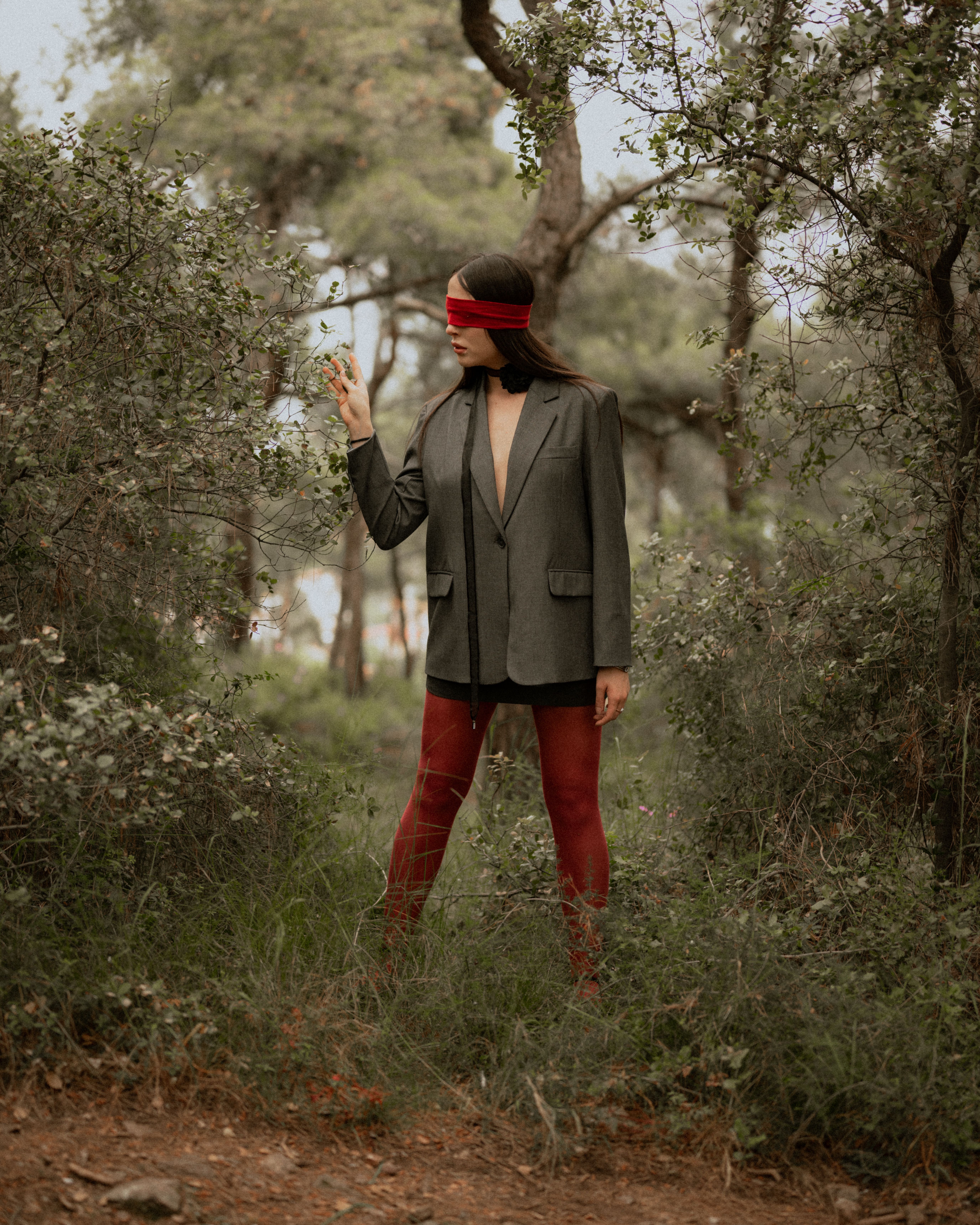 Model in a gray suit and red stockings poses blindfolded in a serene forest setting.
