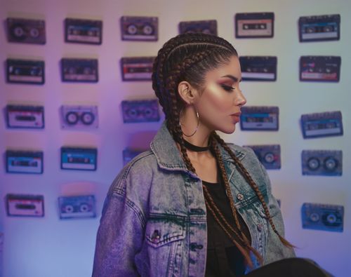 Free Stylish woman with braided hair and denim jacket posing against cassette-covered wall. Stock Photo