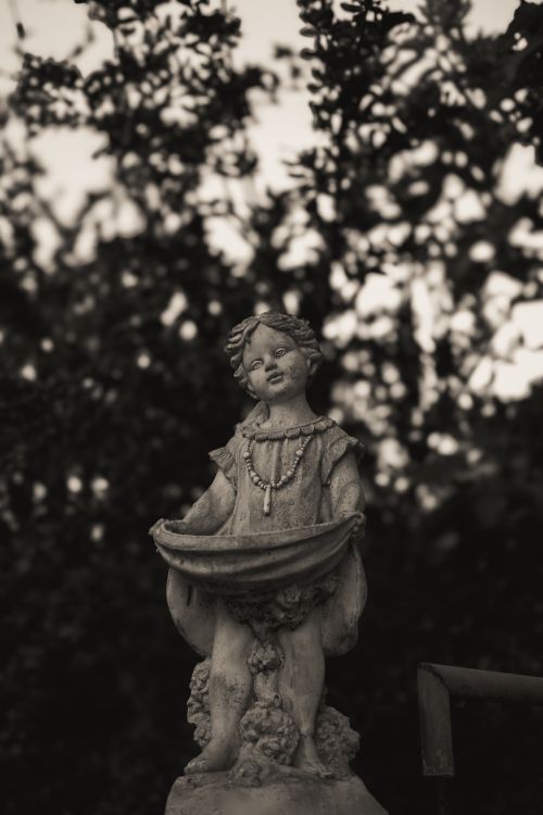 Free A vintage stone statue of a child amidst lush greenery, exuding a timeless charm. Stock Photo