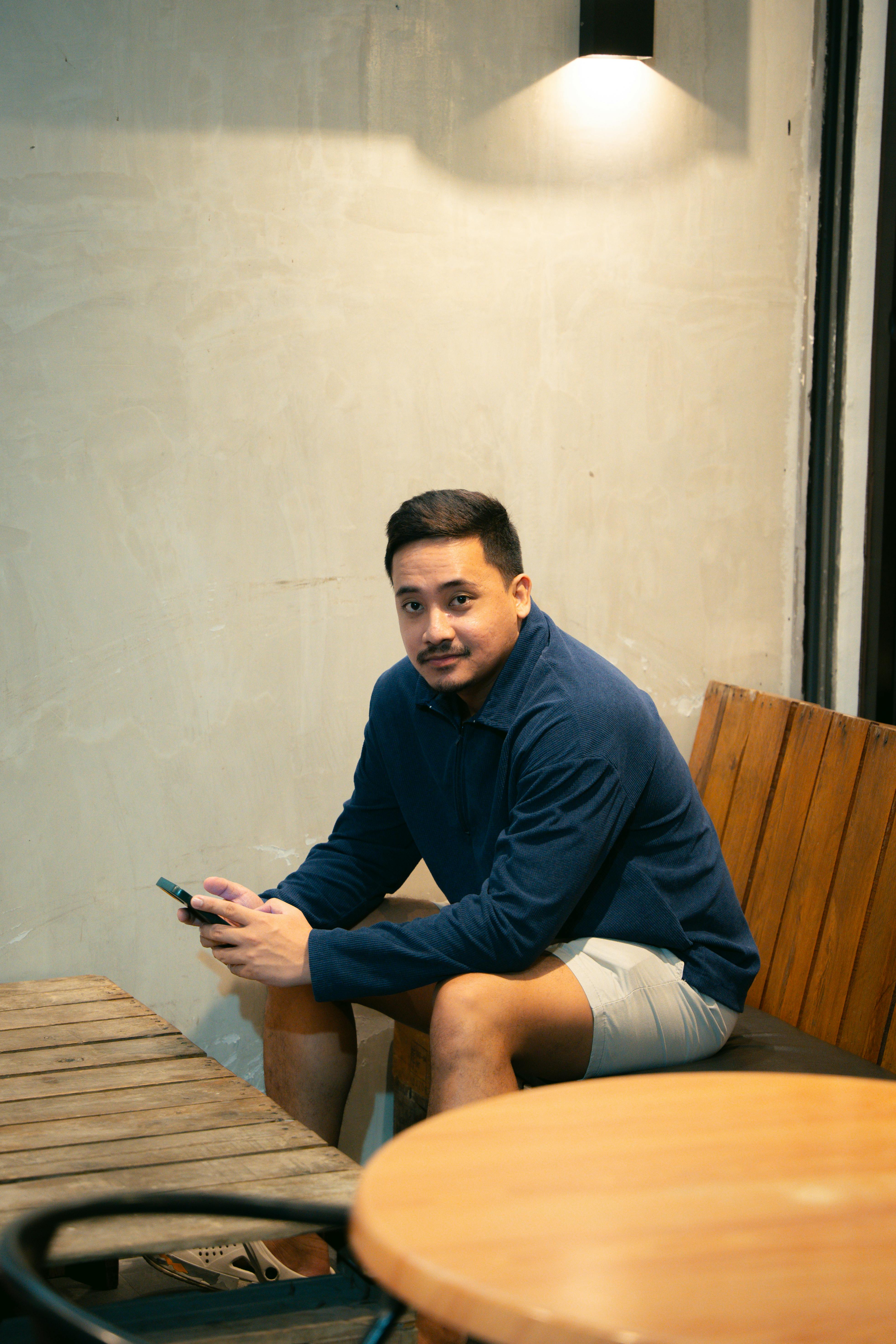 Free Asian man in casual attire sitting and holding a smartphone indoors under soft lighting. Stock Photo