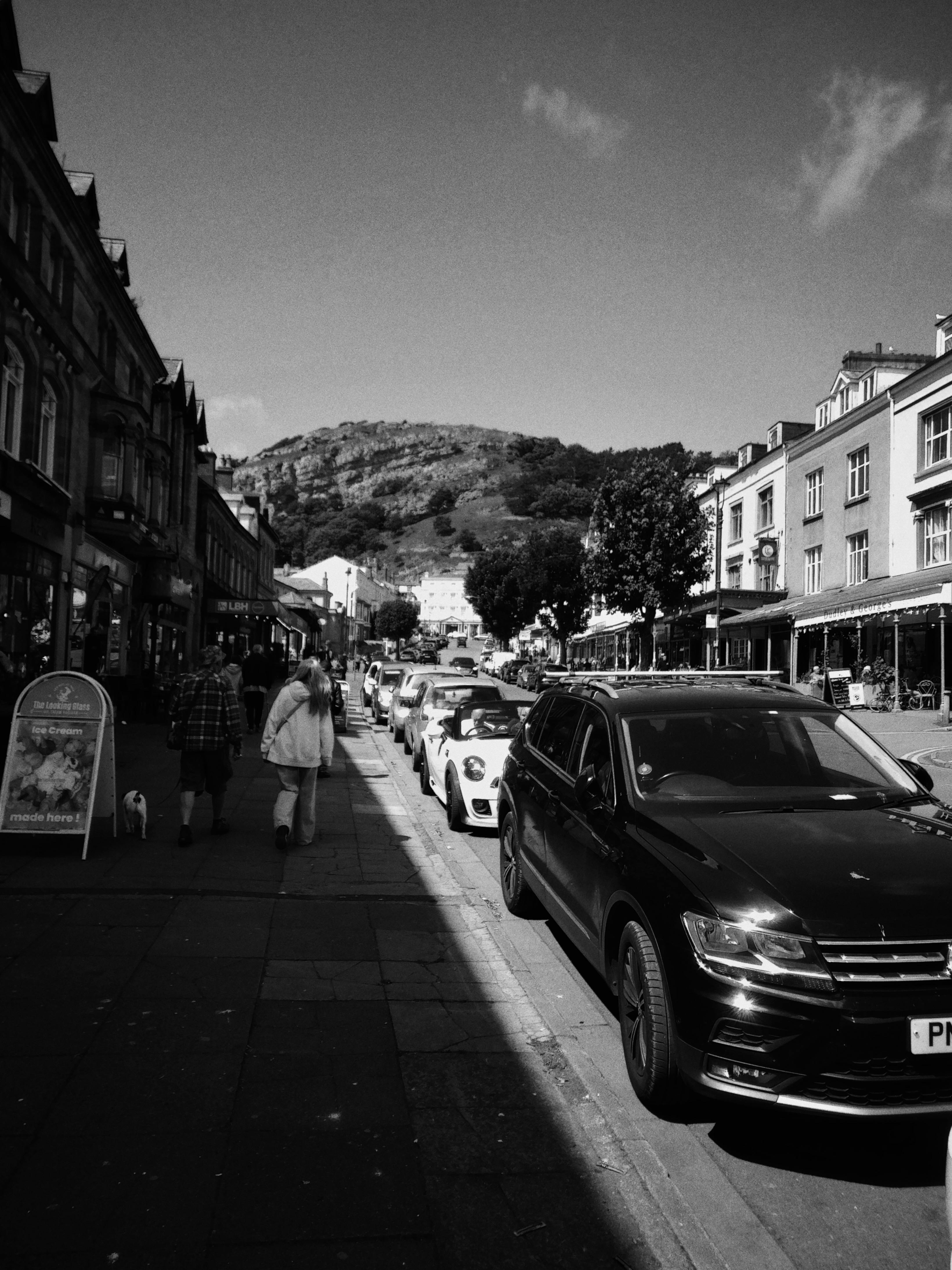 Monochrome view of a bustling street with parked cars and pedestrians set against hills.