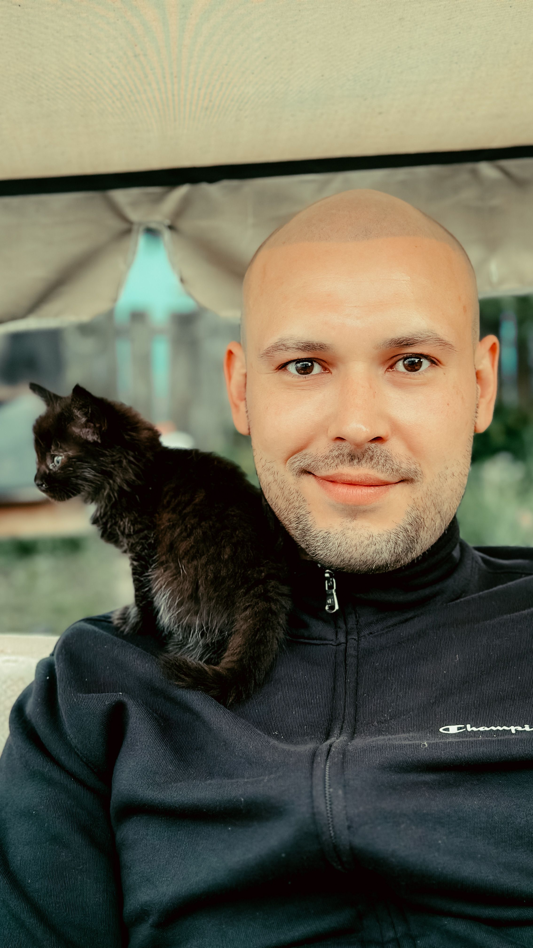 Smiling man with a black kitten on his shoulder, enjoying an outdoor moment.