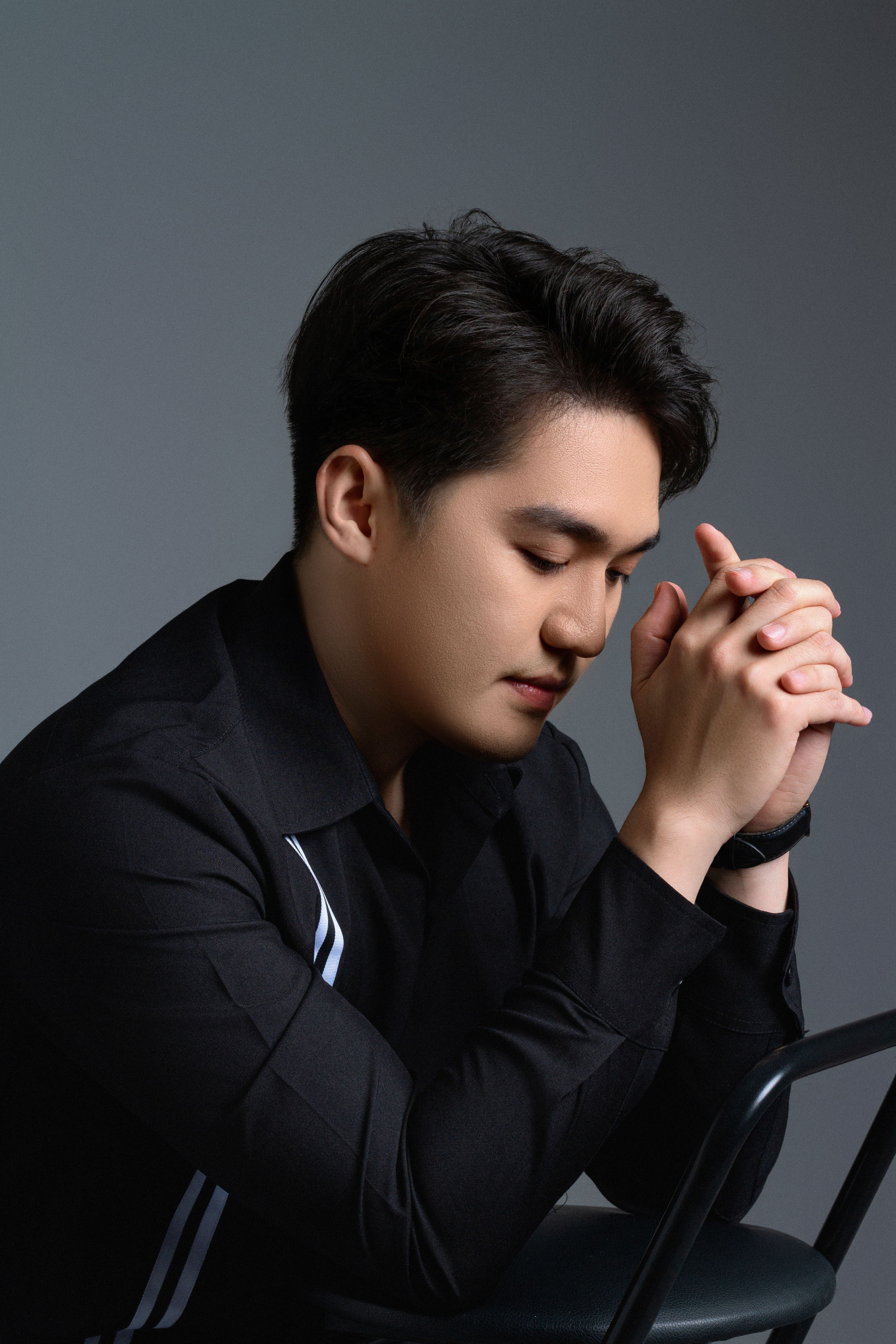 Portrait of an Asian man in deep thought, hands clasped, in a professional studio setting.