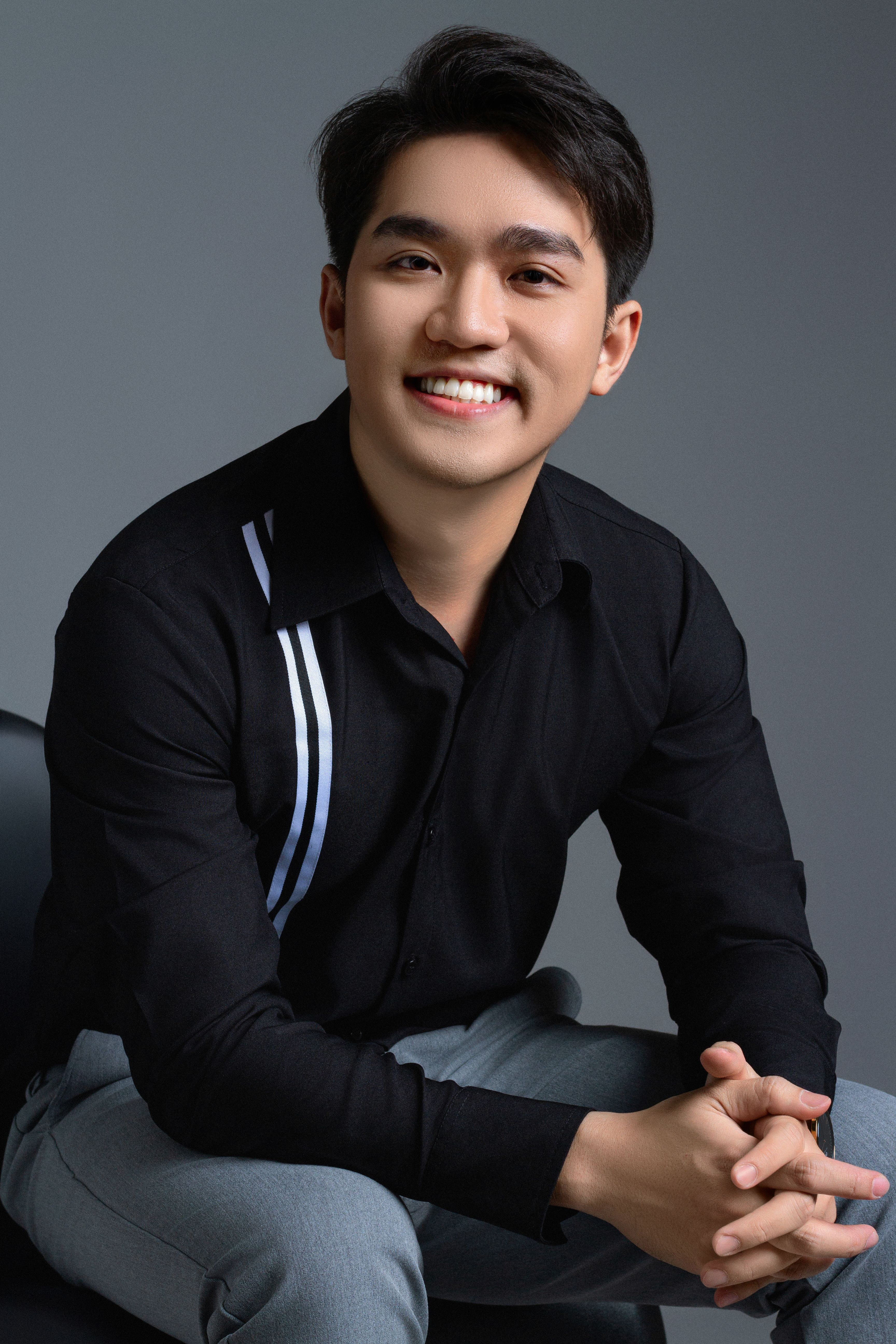 Studio portrait of a smiling young man in smart casual attire sitting against a gray background.