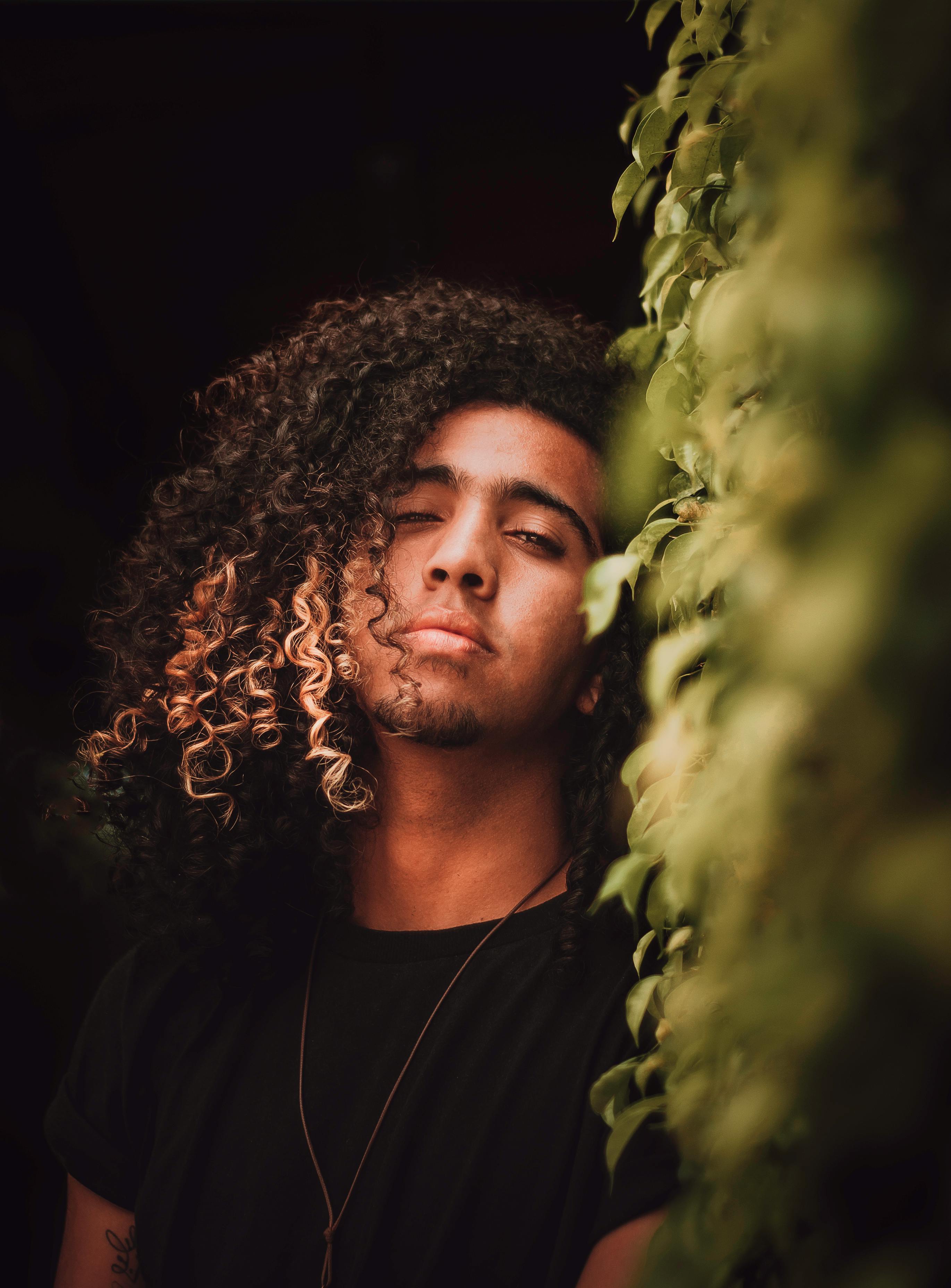 Portrait of a young man with curly hair leaning on a hedge, wearing a black shirt, outdoors.