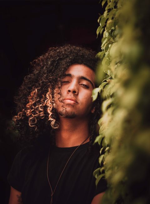 Portrait of a young man with curly hair leaning on a hedge, wearing a black shirt, outdoors.