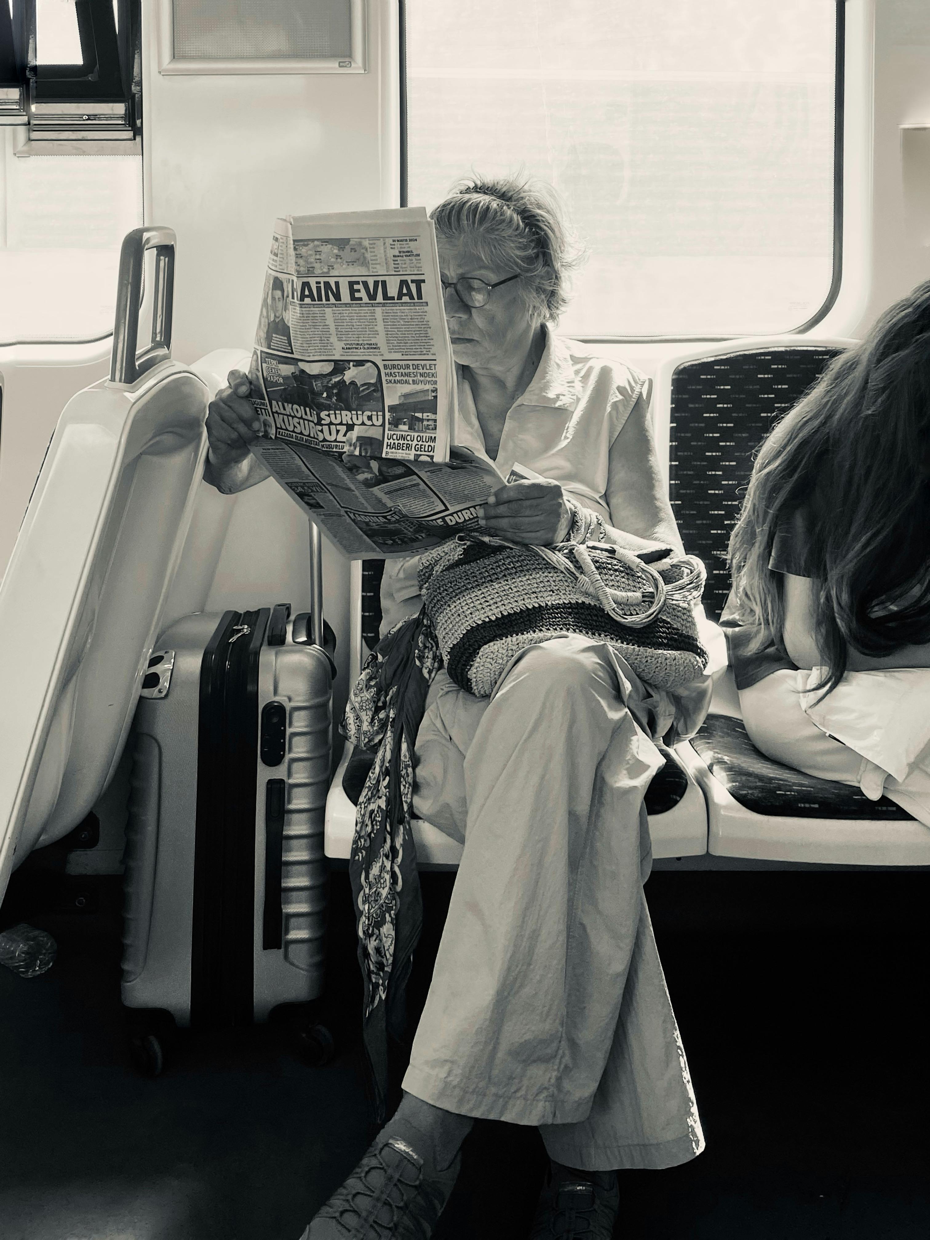 A senior woman reads a newspaper while traveling on a train, next to luggage.