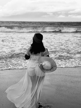 A woman in a white dress holding a sunhat stands facing the ocean waves.