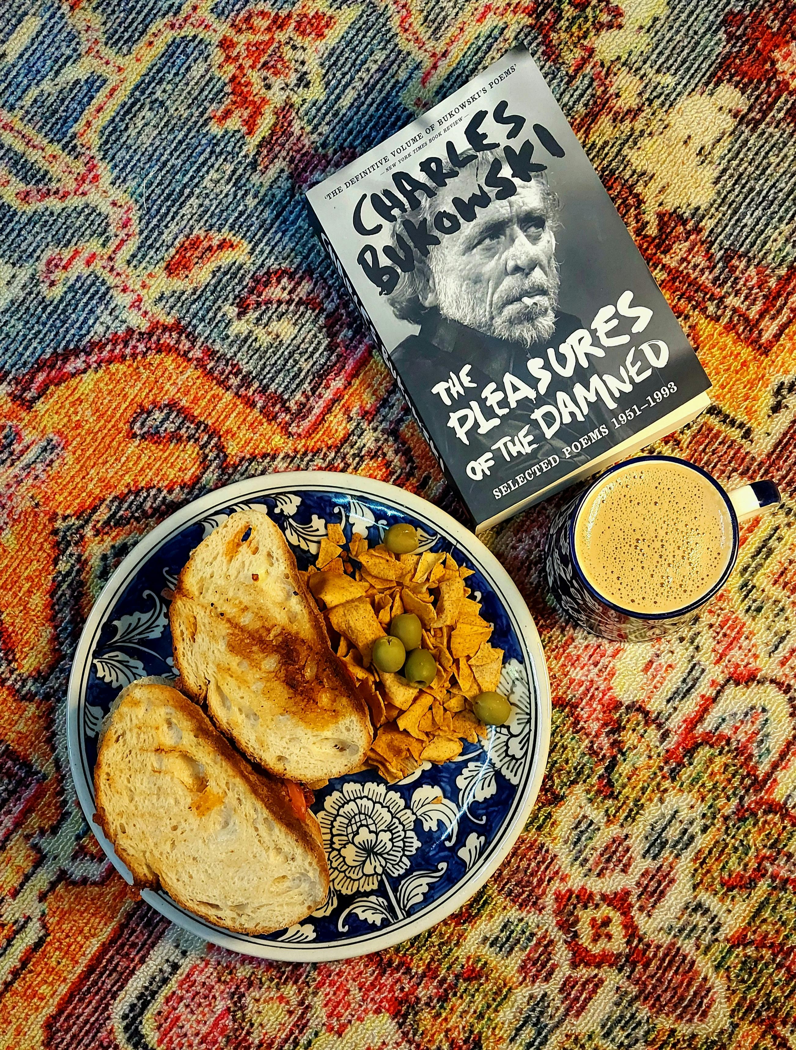 Top view of a breakfast setup with toast, coffee, and a Charles Bukowski book on a patterned rug.