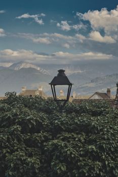 A classic lamppost surrounded by lush greenery with a mountain backdrop, under a cloudy sky.