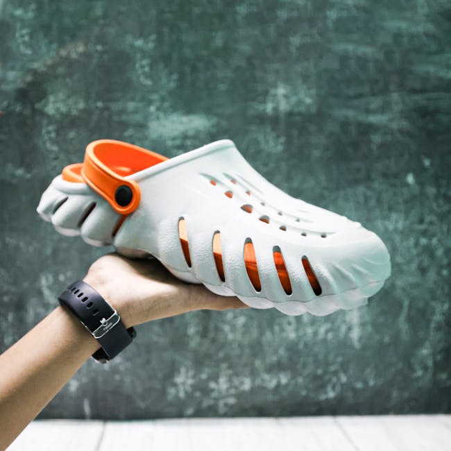 Closeup of a hand holding a stylish white and orange clog shoe against a textured background