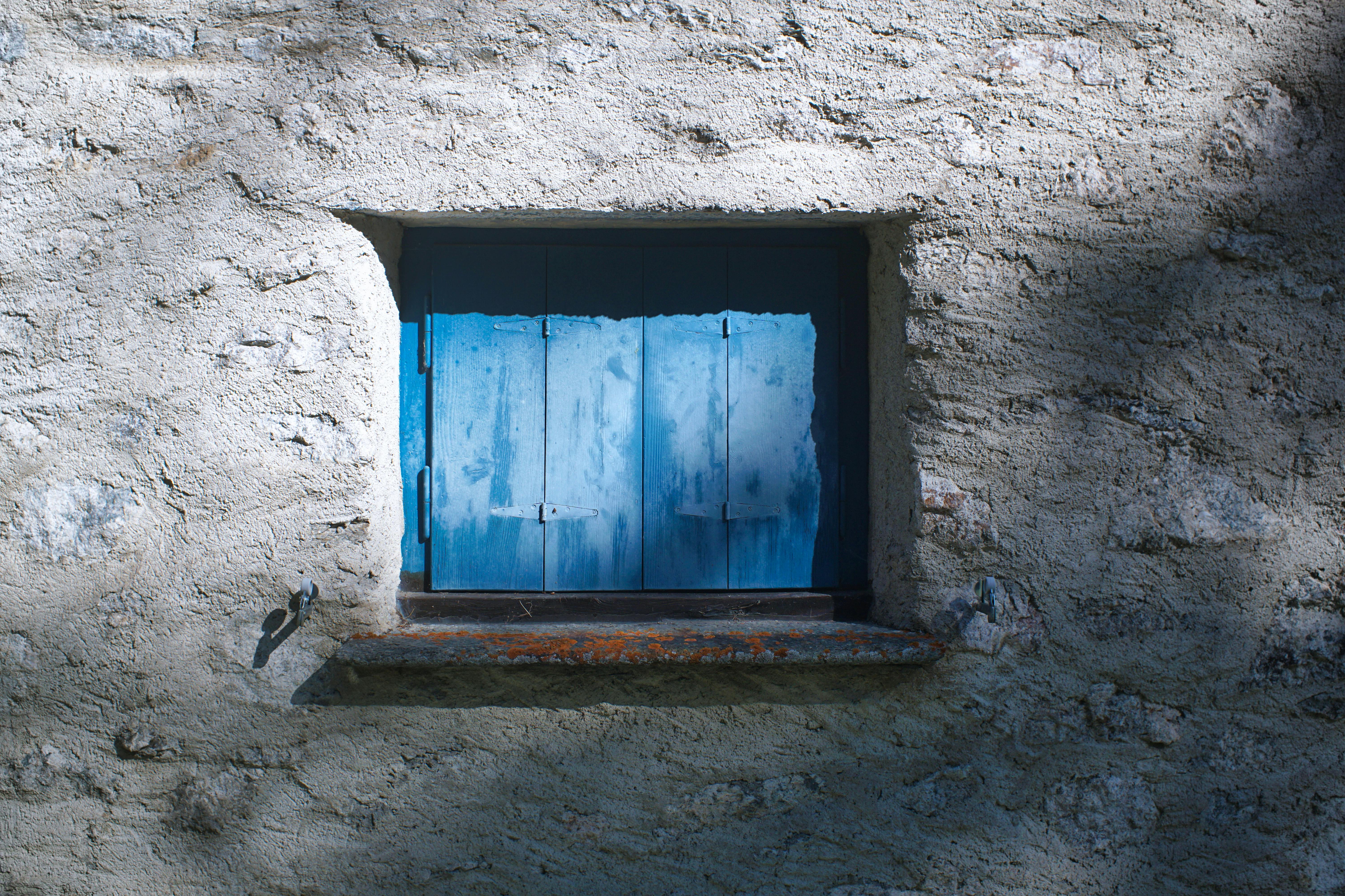 A rustic blue shutter on an old stone building exterior illuminated by sunlight.