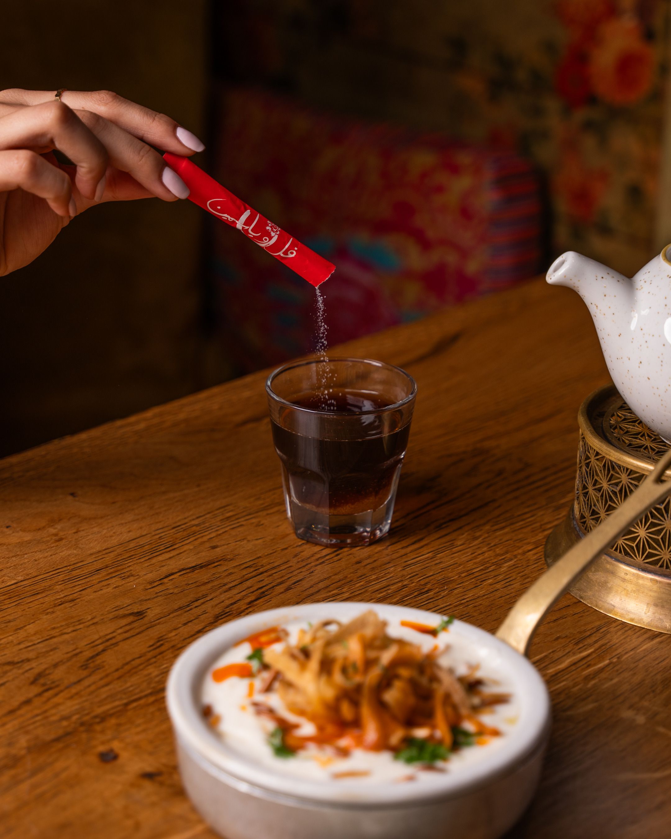 A hand pouring sugar into a glass of tea on a wooden table next to a plate of food.