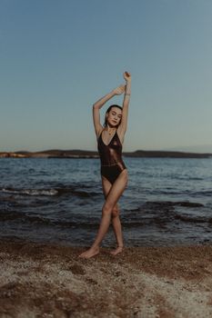 Elegant woman in swimsuit posing on a serene beach during sunset, arms raised.