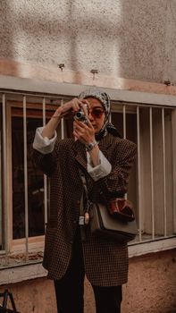 Stylish woman wearing hijab and sunglasses captures a photo outdoors with vintage camera.