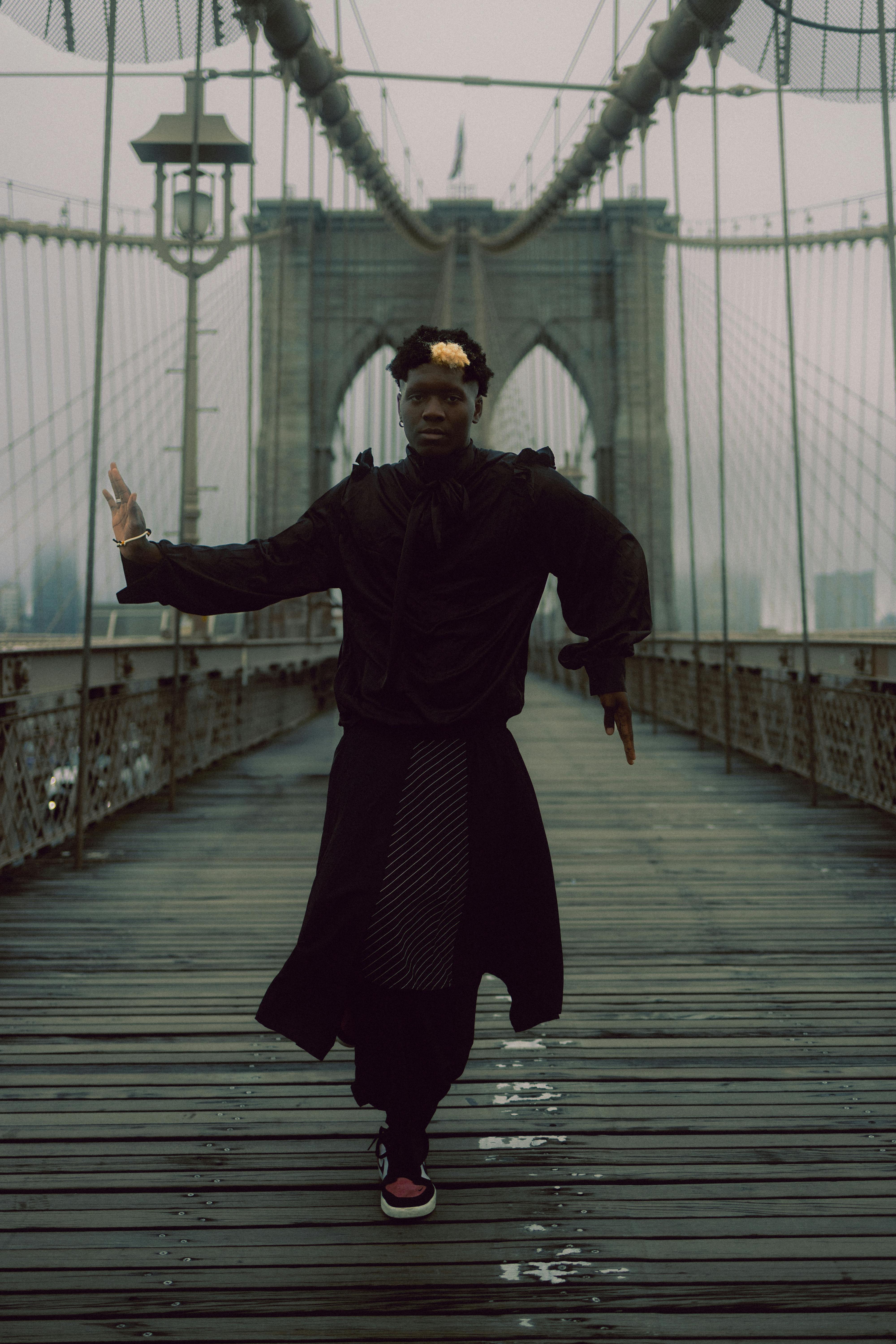 Young man wearing black clothing poses dramatically on the iconic Brooklyn Bridge amid fog.