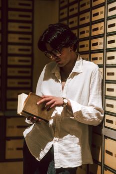Young man with glasses reading a book in an archive room with vintage drawers.