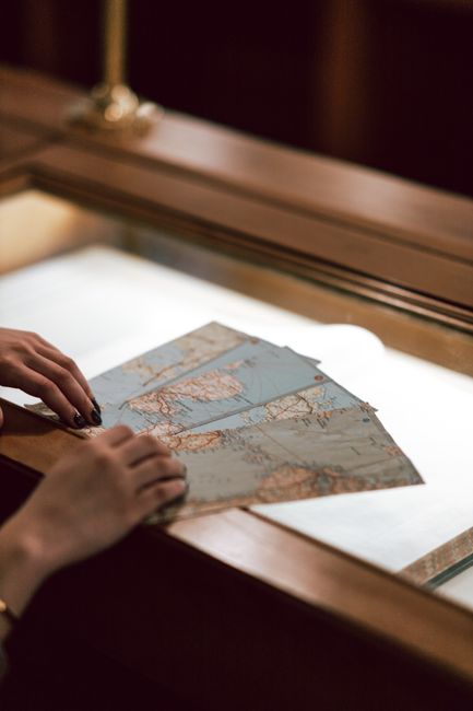 A close-up of a woman's hands holding vintage maps on an illuminated glass table in a dim room.