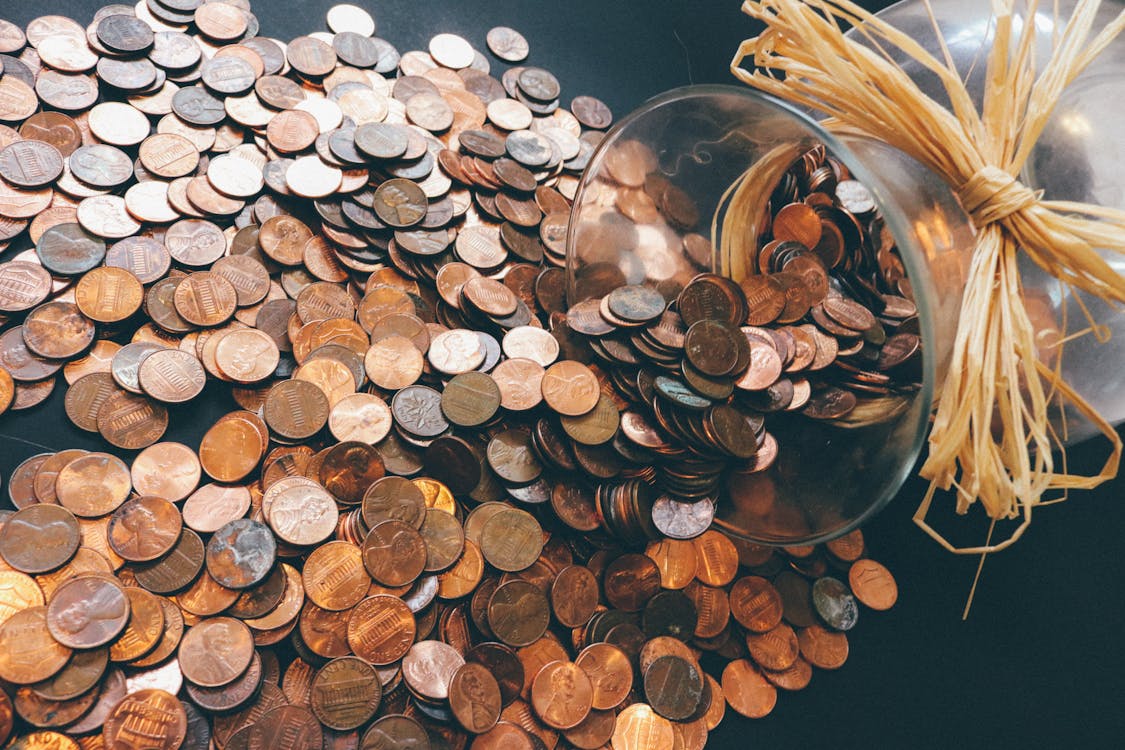 An overturned clear glass jar spilling an abundance of shiny and tarnished pennies onto a dark surface, reflecting the concept of savings and expenditures. A raffia ribbon is tied around the neck of the jar, adding a rustic touch to the image of scattered coins.