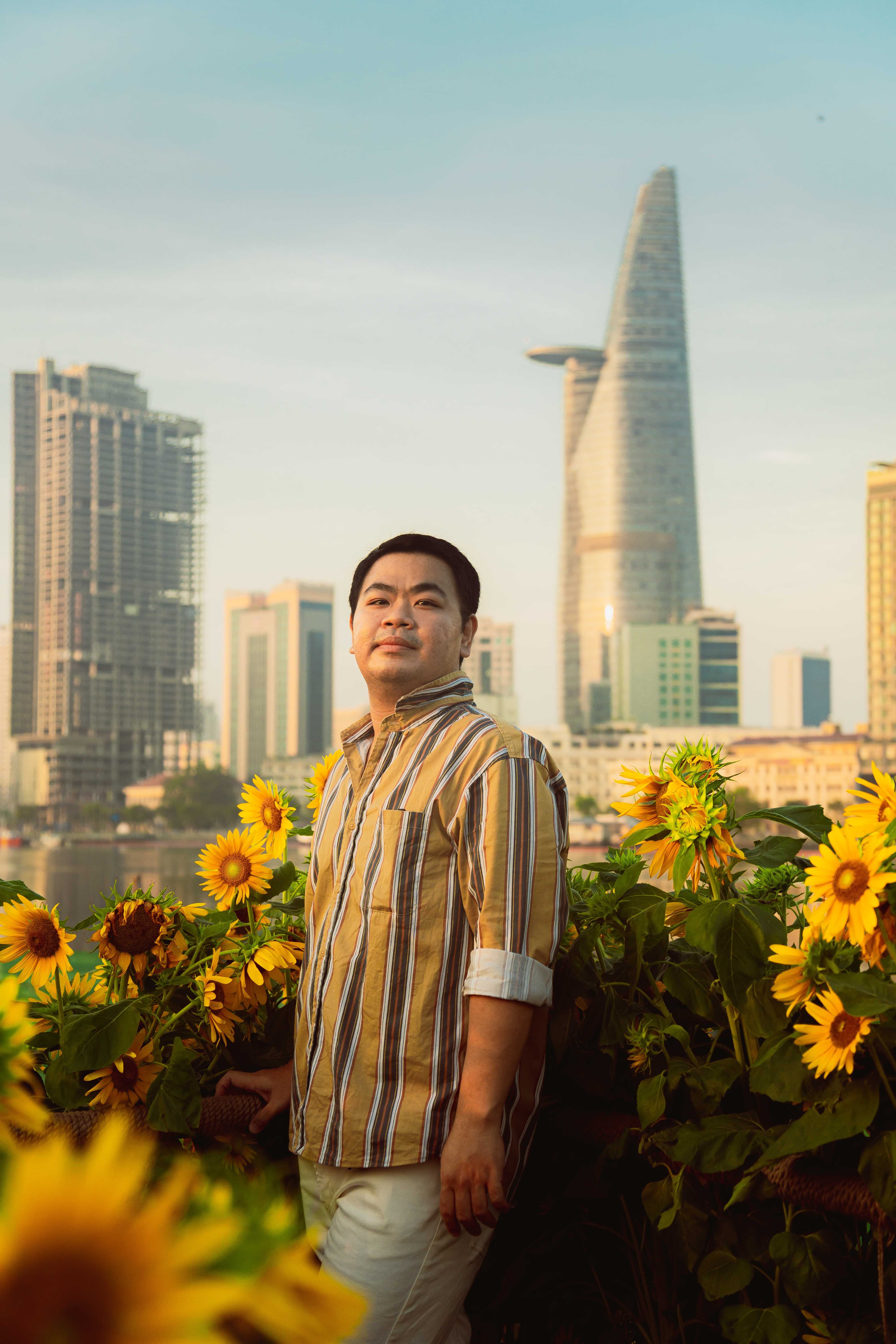 A man stands among sunflowers with the Ho Chi Minh City skyline in the background at sunset.