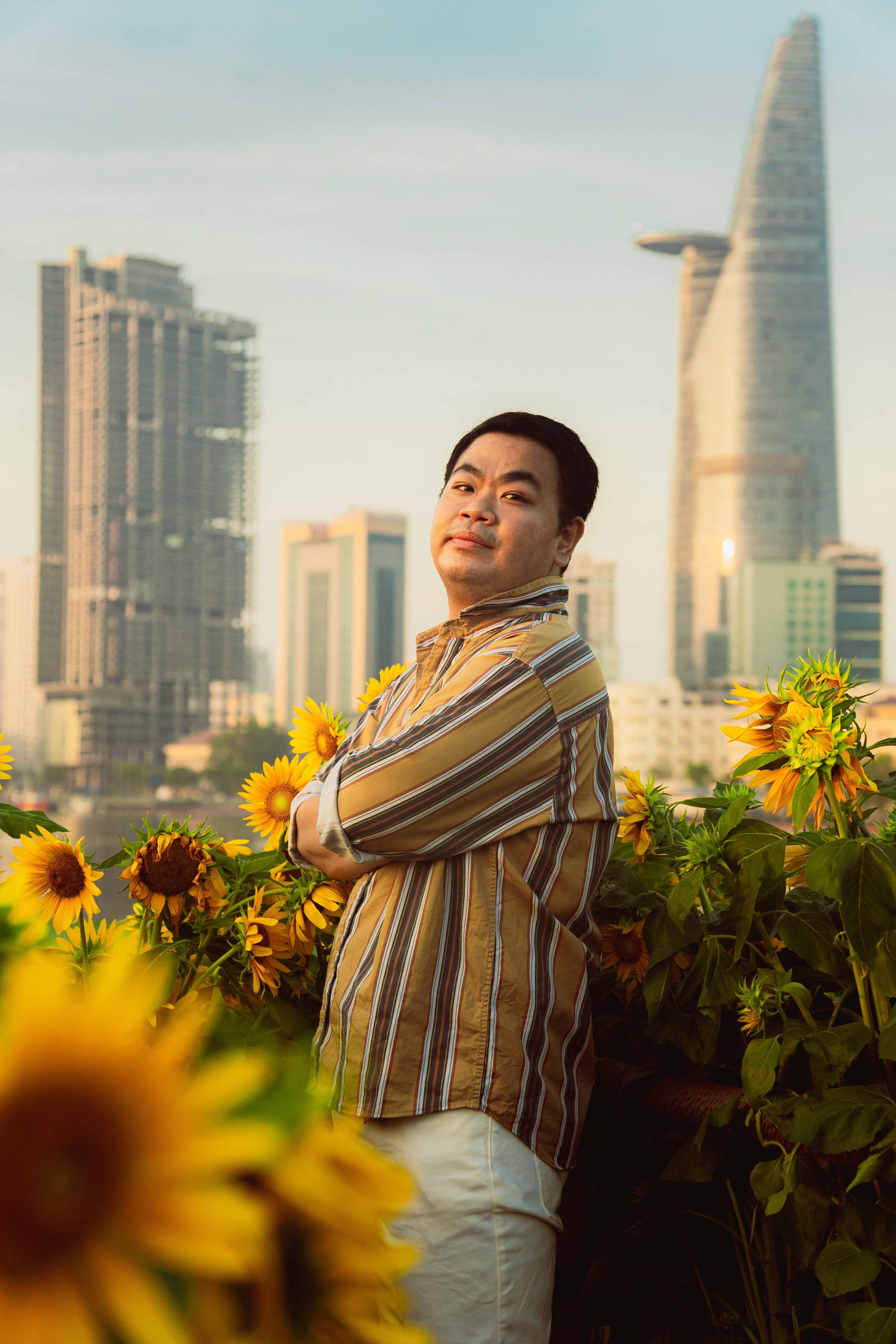 A man stands in a sunflower field with Ho Chi Minh City skyline in the background during sunset.