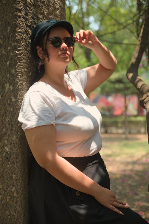 A woman in a white shirt and black skirt leaning against a tree