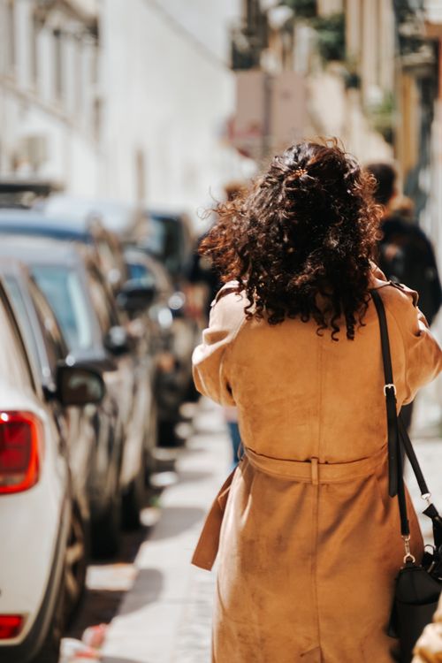 Free Brunette woman in a coat walking along a city sidewalk with cars parked beside. Stock Photo
