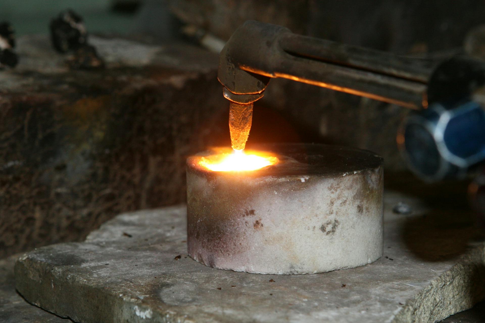 A close-up view of a metal smelting process using a torch, illustrating heat and sparks.