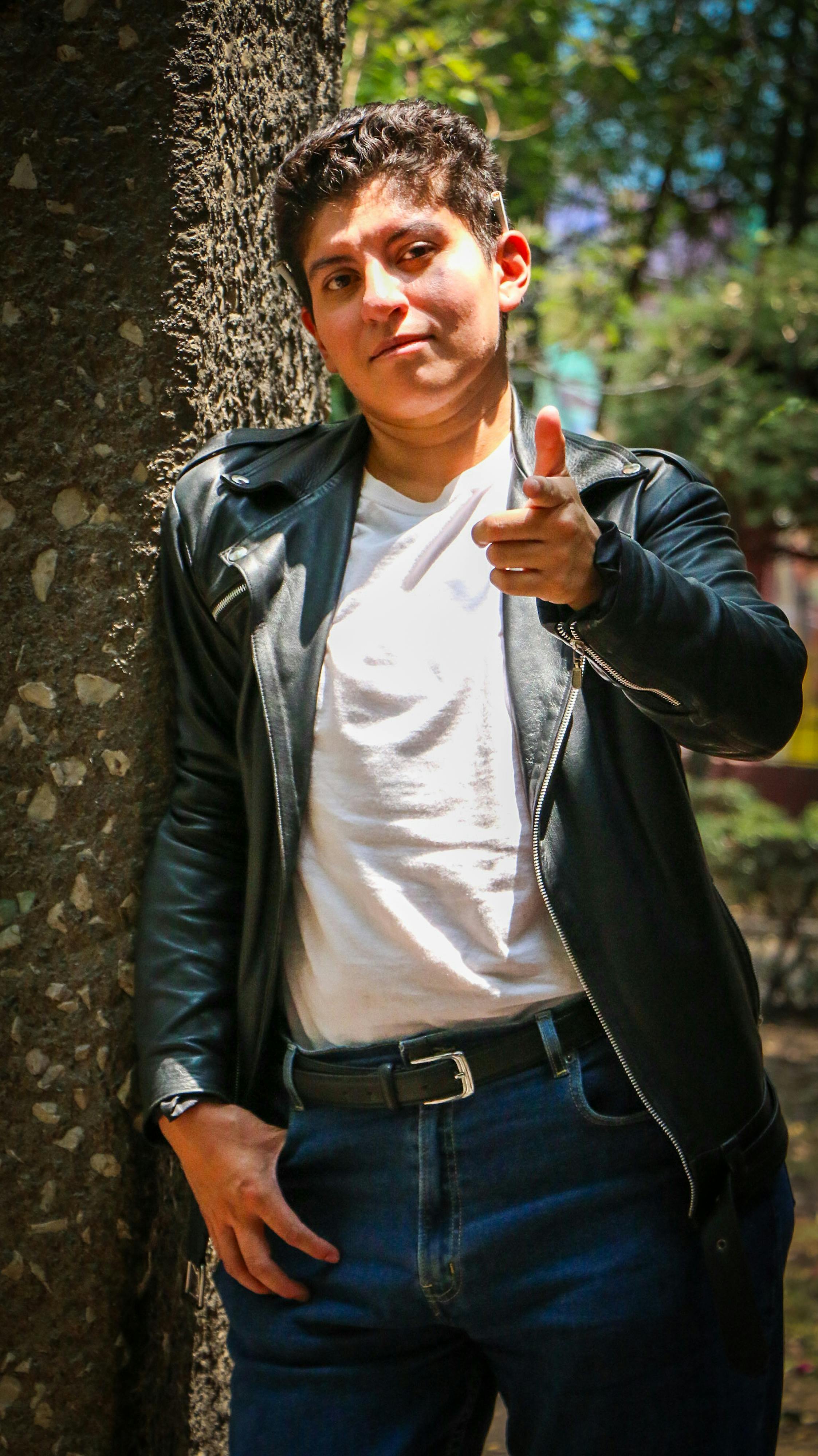 A young man in a leather jacket casually leans against a tree in a sunlit park.