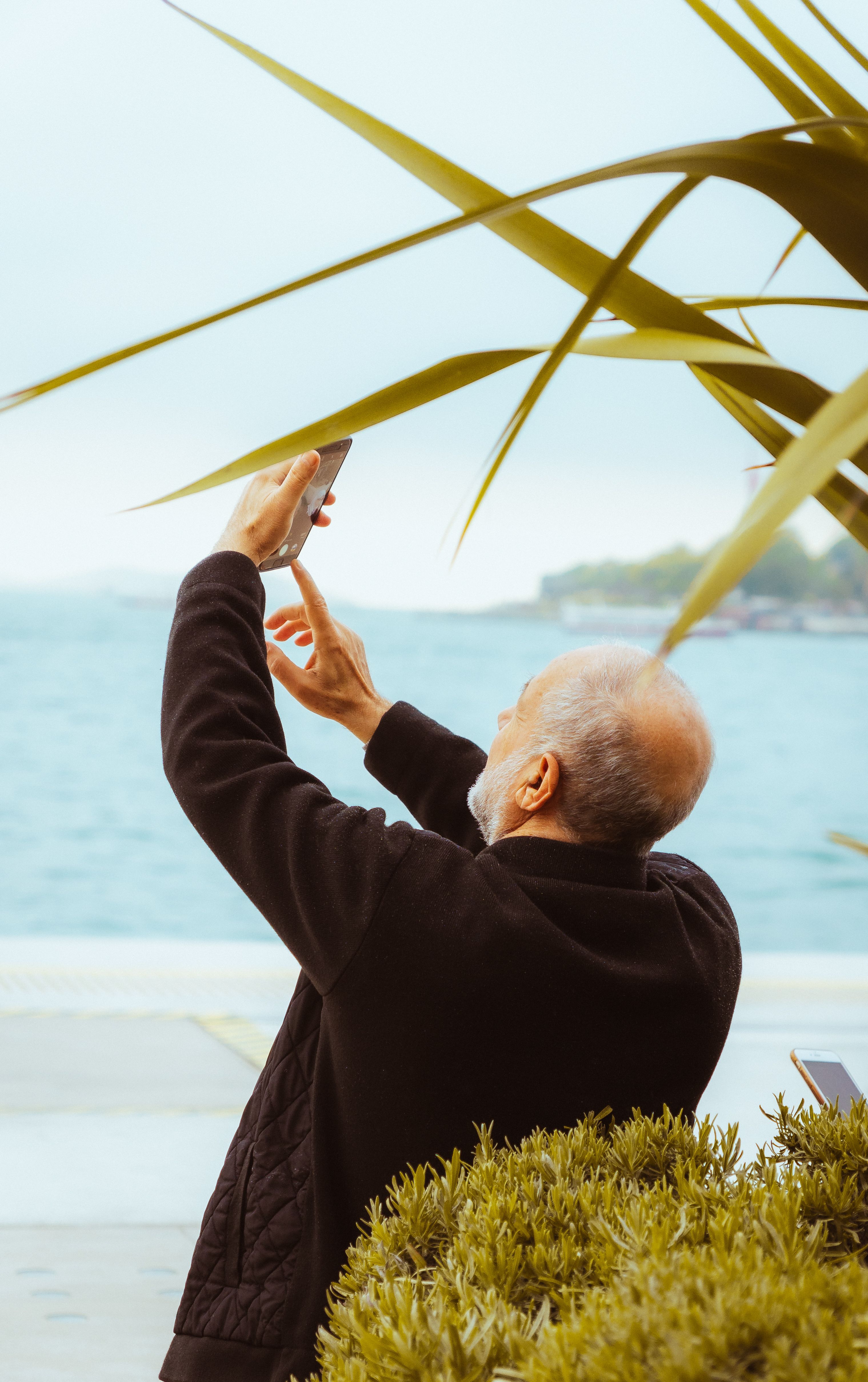 Elderly man captures the scenic İstanbul coast using a smartphone, showcasing vibrant outdoor life.