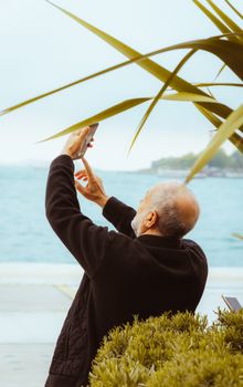 Elderly man captures the scenic İstanbul coast using a smartphone, showcasing vibrant outdoor life.