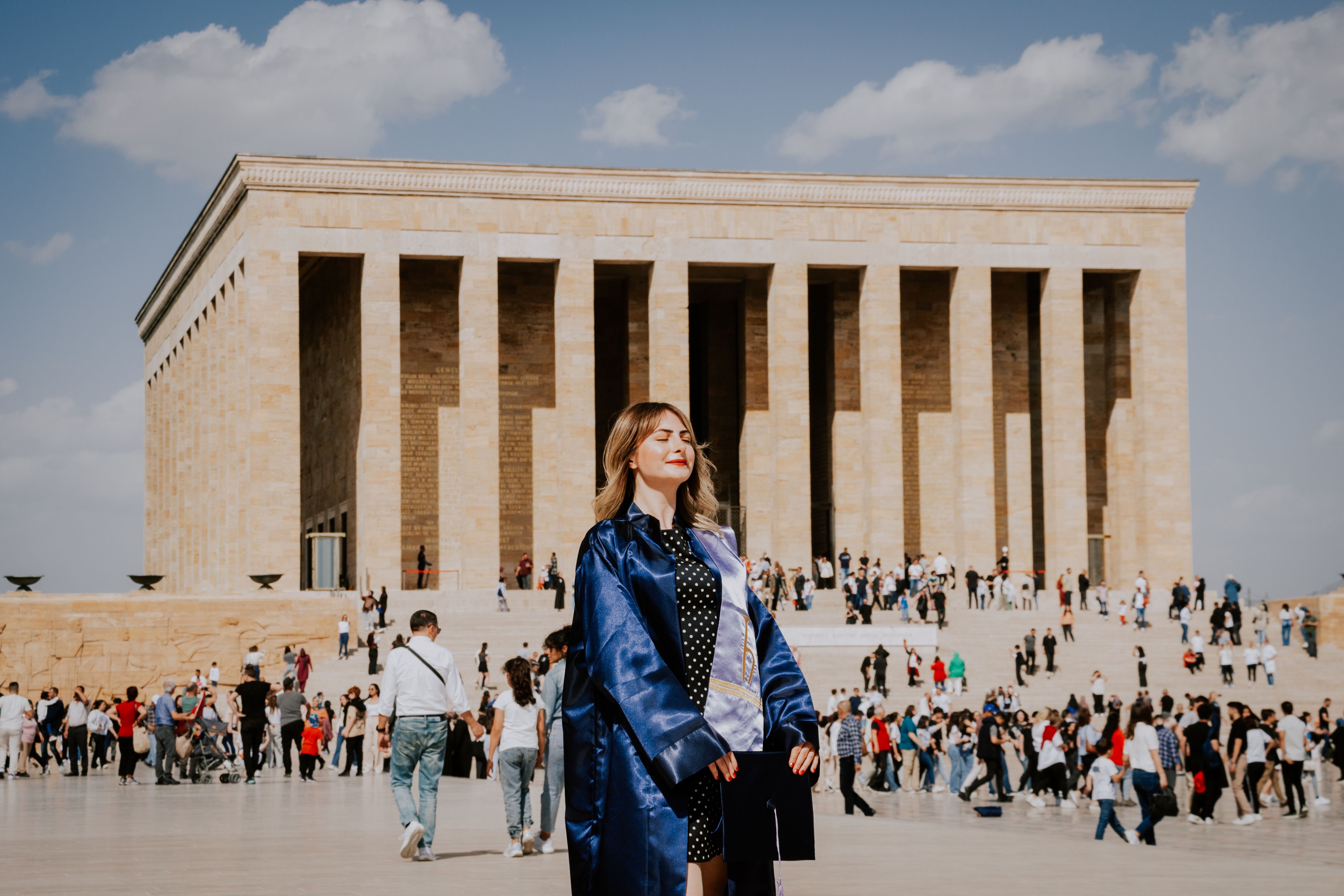Woman in graduation gown at Anitkabir, surrounded by tourists on a sunny day.