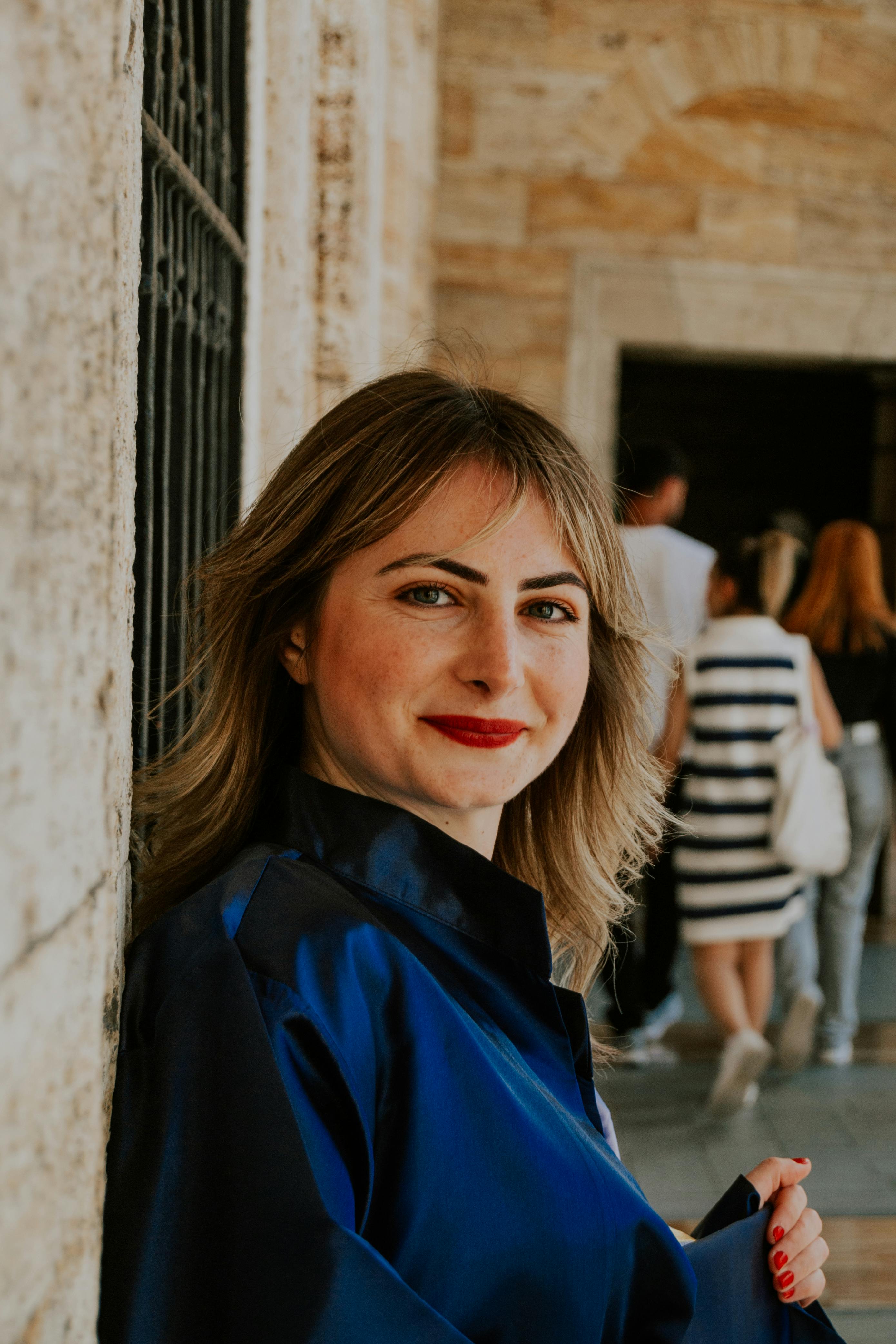 Smiling woman in a blue shirt leaning against a stone wall outdoors.