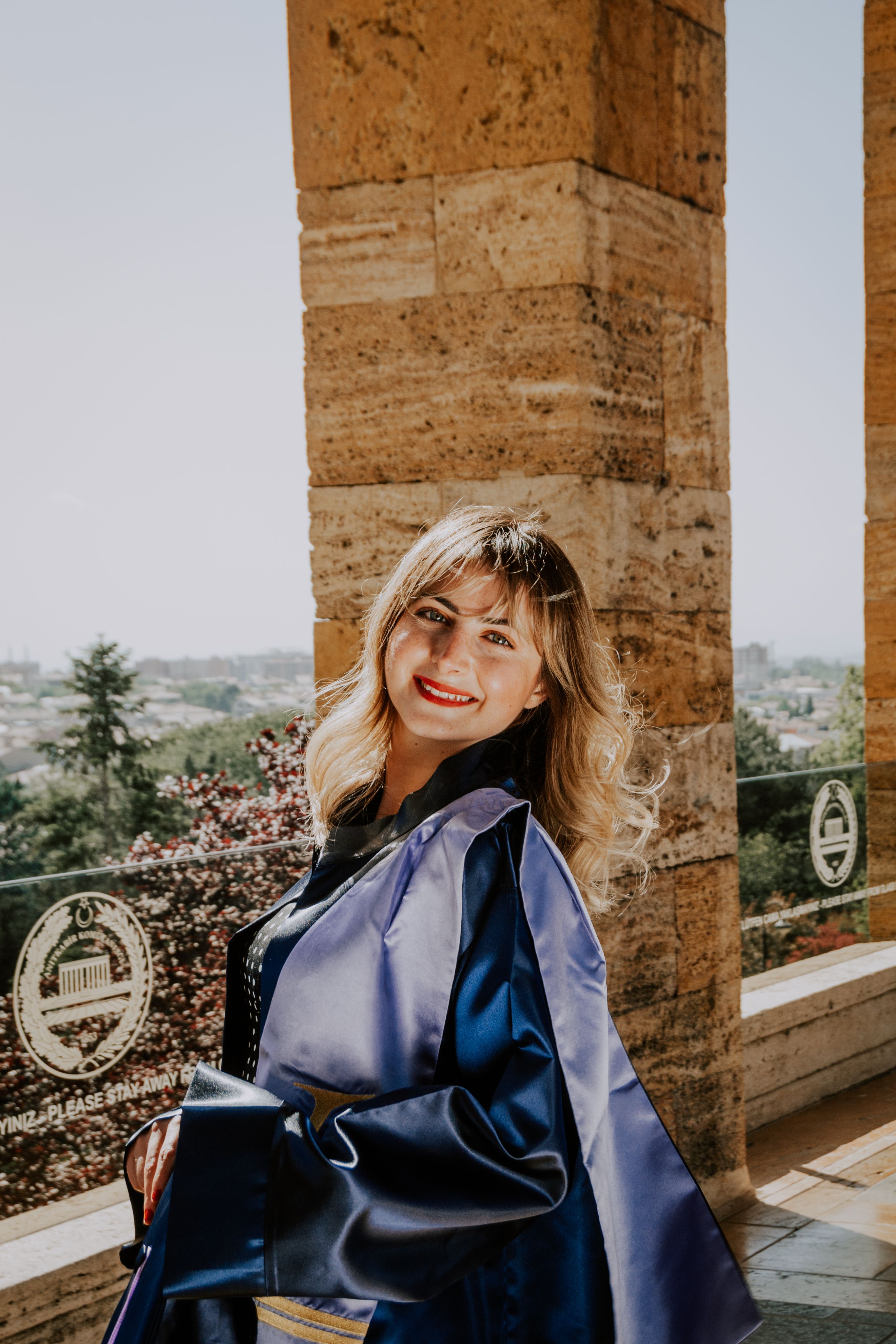 A smiling blonde woman in a graduation gown posing outdoors in a classic setting.