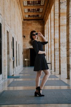 Blonde woman posing stylishly in a black polka dot dress and sunglasses in a stone corridor.