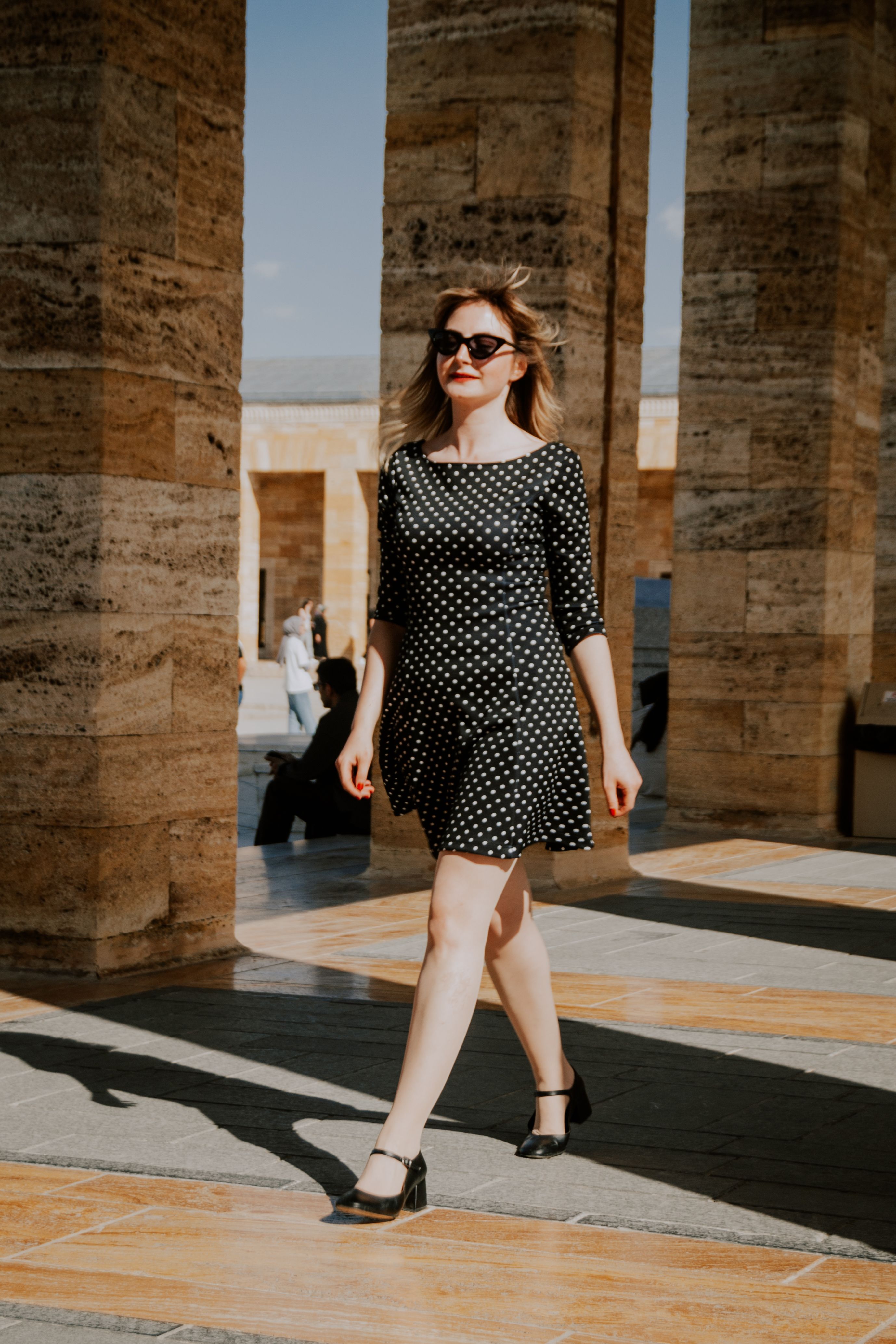 Mulher elegante em vestido de poás caminha com confiança ao ar livre entre colunas de pedra.