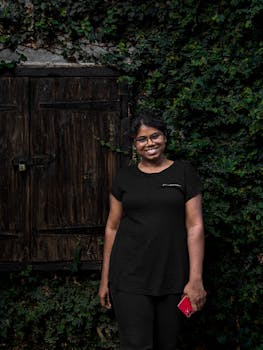 Woman with a smartphone posing happily in front of a rustic wooden shed, surrounded by greenery.