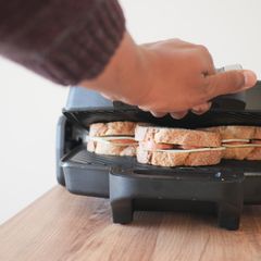 Hand pressing the lid of a black sandwich grill down on two thick sandwiches, on a light wooden worktop