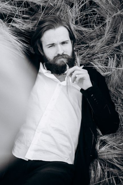 Monochrome portrait of a bearded man in a suit, lying in a field, conveying a relaxed yet serious vibe.