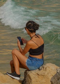 Woman sits on rocky shore in Benidorm, Spain, enjoying summer sea views while using smartphone.