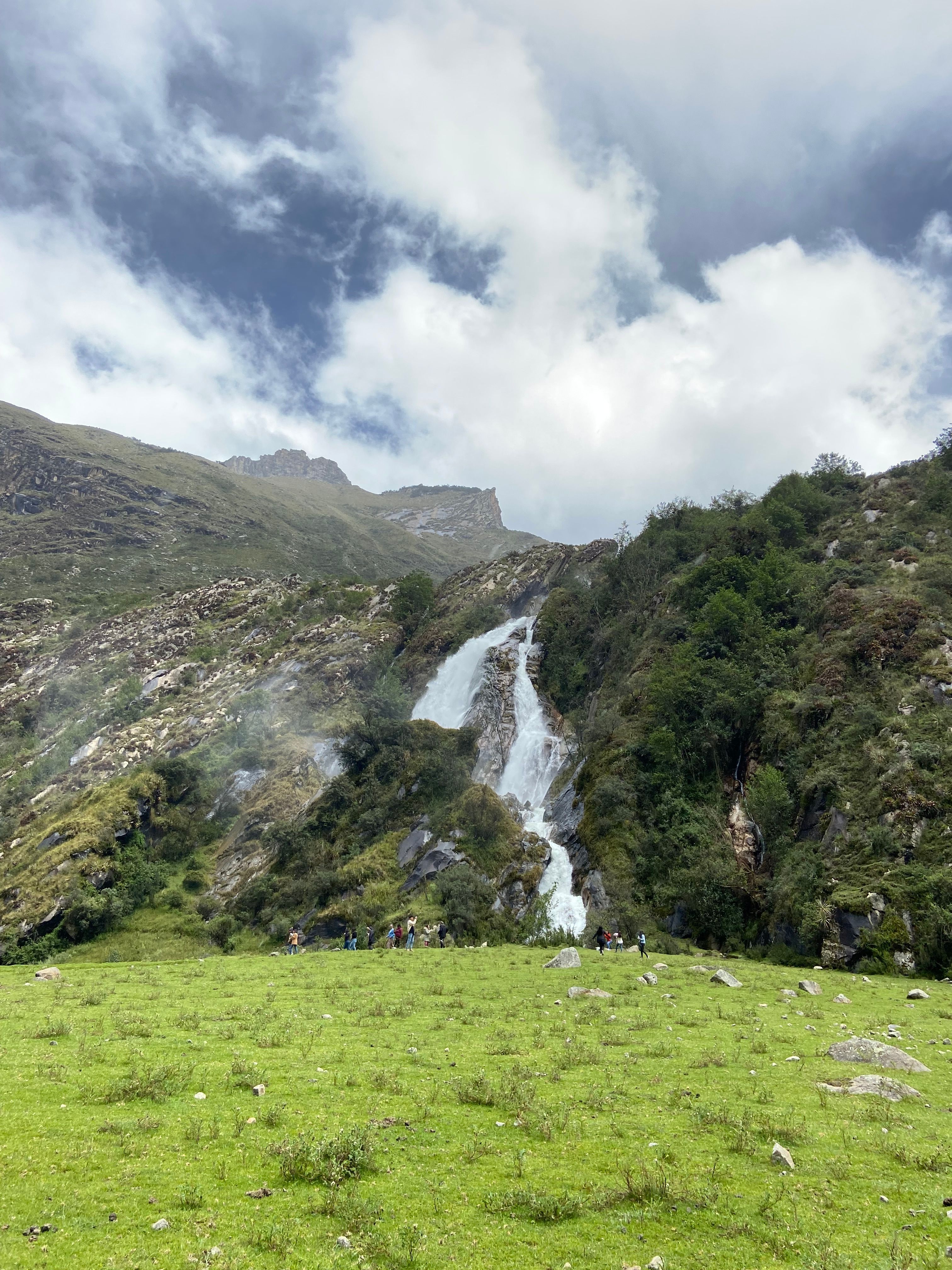 Stunning mountain waterfall cascading into lush green meadow under cloudy sky.