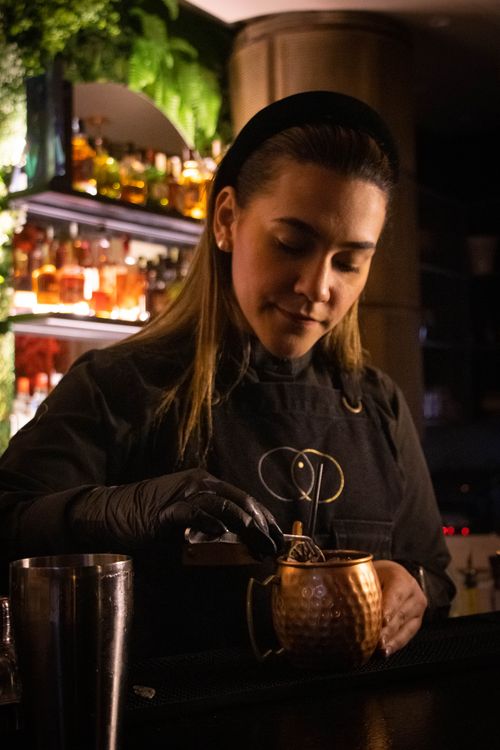 Free Female bartender preparing a cocktail in a dimly lit bar with bottles in the background. Stock Photo