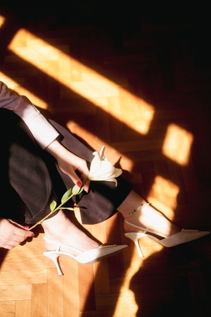 A woman in white heels and black pants holds a white rose, sitting in a sunlit room with a parquet floor.