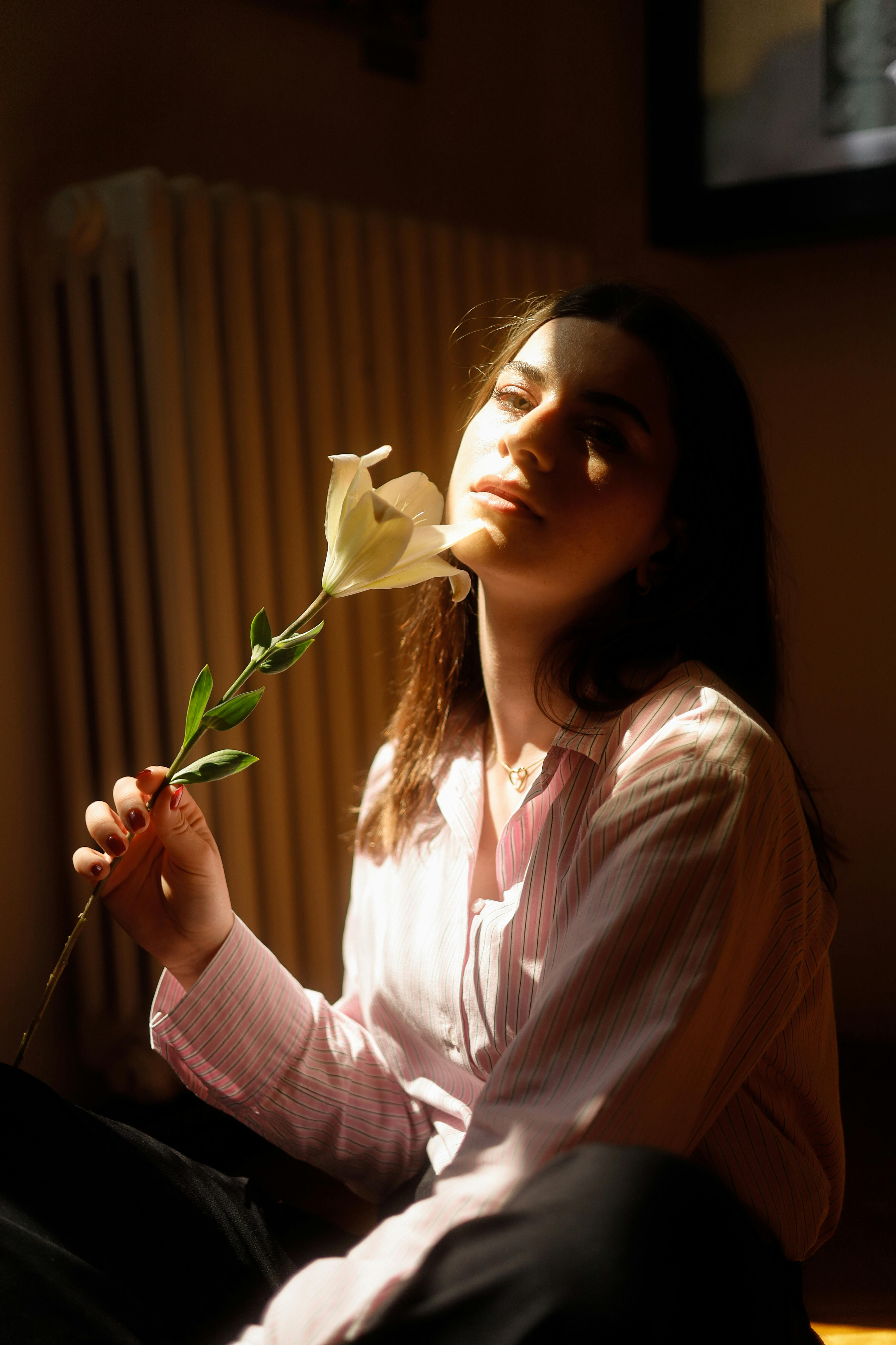 A serene woman with long hair holding a white flower, bathed in soft sunlight indoors.