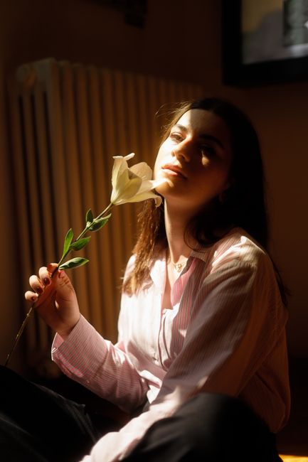 A serene woman with long hair holding a white flower, bathed in soft sunlight indoors.