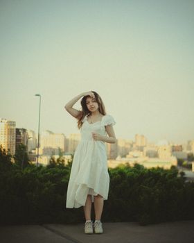 A young woman in a white dress poses thoughtfully against a scenic urban backdrop.