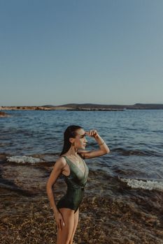 A woman in a bathing suit stands on a rocky beach, looking at the horizon on a sunny day.